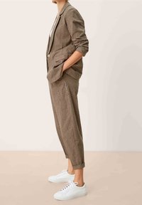 Olijfgroen geperforeerd stoffen pak met een op maat gemaakte blazer en taps toelopende broek, gecombineerd met witte sneakers; heeft een gestructureerd ontwerp en zakken.
