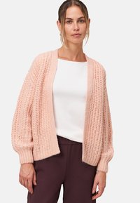 Lichtroze, gebreide cardigan met een losse pasvorm, geribbelde textuur en lange, ballonmouwen. Draagt over een wit top met donkere broek.