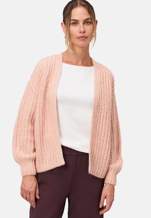 Cardigan tricoté rose clair avec une coupe ample, texture côtelée et manches longues bouffantes. Porté sur un haut blanc avec un pantalon sombre.