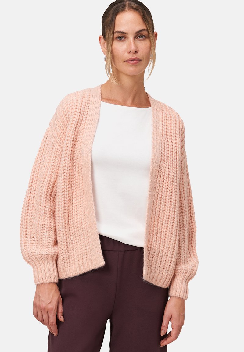 Lichtroze, gebreide cardigan met een losse pasvorm, geribbelde textuur en lange, ballonmouwen. Draagt over een wit top met donkere broek.