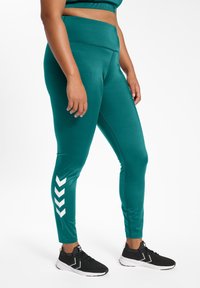 Teal sportleggings met een hoge taille, een strakke textuur en een wit zigzagpatroon op het buitenbeen. Gecombineerd met zwarte sneakers.