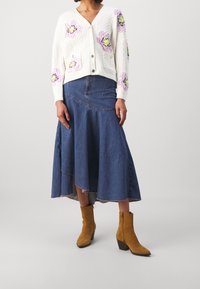 Kobieta ubrana w biały kardigan z fioletowymi kwiatami, asymetryczną niebieską dżinsową spódnicę midi oraz beżowe zamszowe botki na obcasie.