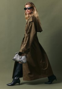 Olīvzaļš trenšmantu ar jostu viduklī, melni ceļgalu augstie norādītie zābaki un pelēka čūsku ādas tekstūras rokassoma.