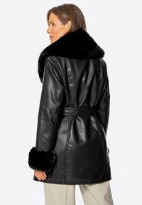 Schwarzer Kunstledermantel mit großem schwarzem Fellkragen und -manschetten. Mit einem taillierten Schnitt und einem Gürtel in der Taille. Glatte Textur, knielang.