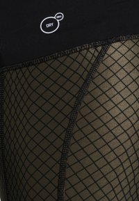 Ropa deportiva negra con una superposición de malla con un patrón de diamantes en verde oliva. Incluye una etiqueta 'DRY' cerca de la cinturilla.