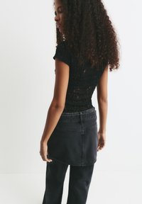 Frau mit lockigem Haar in einem schwarzen Spitzenoberteil und einem geschichteten schwarzen Jeansrock über einer Hose, die vor einem weißen Hintergrund steht.