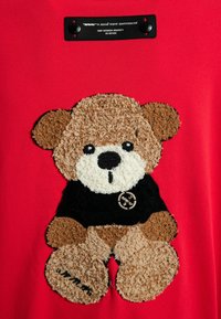 Top de tela roja con un aplique de osito de peluche texturizado en marrón y marrón claro, que lleva una camiseta negra con un logo.
