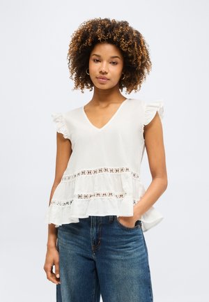 Γυναίκα με σγουρά μαλλιά, που φοράει λευκό αμάνικο μπλουζάκι με δαντελένιες λεπτομέρειες και μπλε τζιν, στέκεται με το ένα χέρι στην τσέπη.