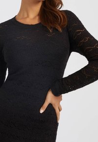 Robe en maille noire à manches longues, avec un motif texturé et une silhouette ajustée. Encolure ronde et bords des manches festonnés.