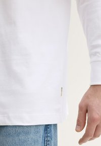 Fehér, hosszú ujjú ing, puha textúrával, oldalsó címkével. Az ing ujjai bordás mandzsettában végződnek, világoskék farmernadrággal párosítva.