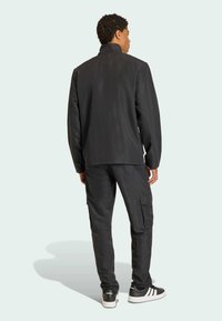 Μαύρο μπουφάν και cargo παντελόνια από ανθεκτικό ύφασμα, με ψηλό γιακά, πλαϊνές τσέπες και άνετη εφαρμογή. Τα υποδήματα είναι μαύρα και λευκά.