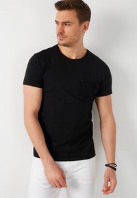 T-shirt nera a maniche corte in cotone con colletto rotondo e taschino sul petto di tessuto testurizzato. Indossata con pantaloni bianchi e un bracciale scuro.