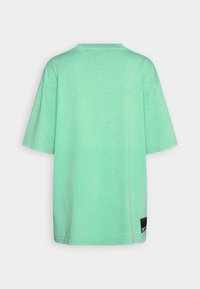 Pudrově zelené oversized tričko s krátkým rukávem, kulatým výstřihem a malou černou obdélníkovou nášivkou poblíž dolního lemu vzadu.