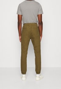 Man in olijfgroene joggerbroek met elastische boorden, een zwart-wit gestreept T-shirt met korte mouwen en witte sneakers, gezien vanuit de achterzijde.