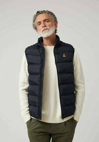 Chaleco acolchado azul marino con cremallera frontal, patrón de costura vertical y parche con logo en el pecho. Lleva puesto encima de un suéter crema y pantalones oliva.