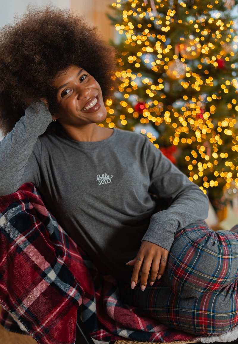 Grijs shirt met lange mouwen met de tekst "Jolly", gecombineerd met een geruite deken en broek in rood en marineblauw, ligt naast een versierde kerstboom.