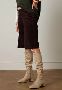 Pantalon court en velours côtelé marron, pull en tricot olive et bottes souples beige montant jusqu'aux genoux avec fermeture éclair. Texture lisse et design structuré.