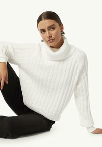 Weißer Strickpullover mit hohem, geripptem Rollkragen, strukturiertem Kabelmuster und lockerer Passform. Kombiniert mit schwarzen, schmal geschnittenen Hosen.