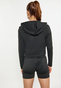 Zwarte cropped zip-up hoodie met capuchon, lange mouwen en een getailleerd ontwerp. Gecombineerd met zwarte, strak zittende shorts, beide van gladde stof.