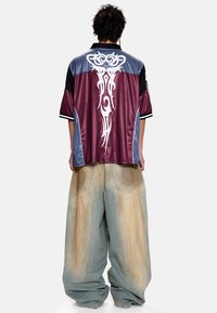 Chemise à manches courtes à panneaux bicolores rouge bordeaux, bleu et noir ; motifs tribaux blancs au dos. Pantalon en jean ample avec finition délavée.