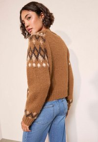 Lühikeste lokkis juustega naine, kes kannab suuri ümmarguseid kõrvarõngaid, pruunikat mustriga kudumisel särki ja siniseid teksapükse, vaatab üle oma õla.