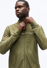 Homme portant une veste zippée vert olive, en train de fermer la fermeture éclair, regardant vers la gauche, sur un fond blanc uni.