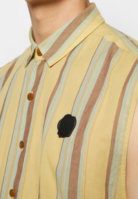 Chemise sans manches à col avec des rayures verticales jaunes, marron et bleues, boutons en bois et un écusson rond noir sur la poitrine.