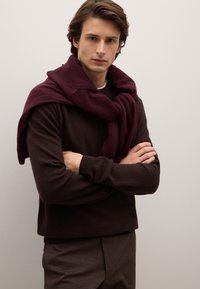 Maglione di lana marrone sovrapposto a una sciarpa bordeaux appoggiata sulle spalle. Il modello posa con le braccia incrociate davanti a uno sfondo neutro.