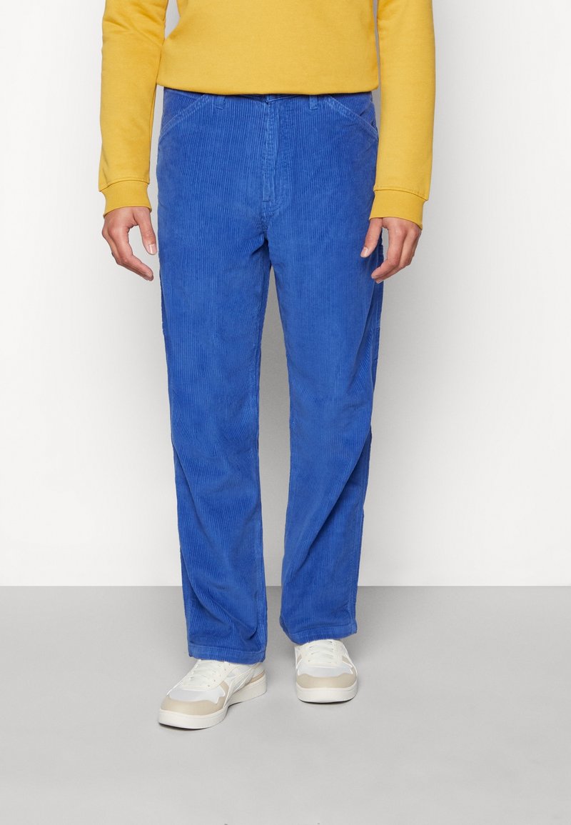 Personne portant un pantalon en velours côtelé bleu vif, un haut à manches longues jaune et des baskets blanches, debout devant un fond uni.