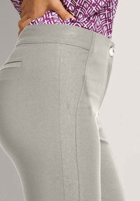 Frau in hellgrauen, figurbetonten Hosen und einer lila gemusterten, in die Hose gesteckten Bluse, mit einer Hand auf der Hüfte.