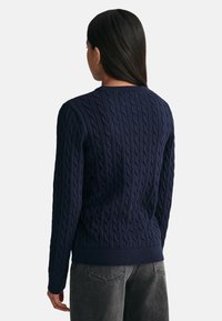Maglione blu navy a trecce con maniche lunghe, polsi e orlo a coste. Indossato sopra jeans grigi, caratterizzati da un motivo testurizzato e in rilievo.