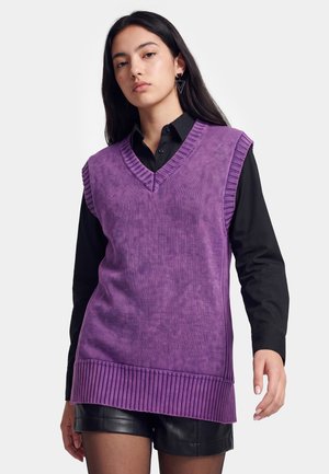 Ung kvinde iført en lilla ærmeløs sweatervest over en sort skjorte med knapper og sorte lædershorts, stående mod en hvid baggrund.