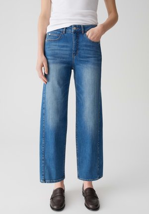 Blauwe wijde jeans van denim hebben een vervaagde afwerking, voorzakken en een knoopsluiting, gedragen met donkere loafers en een witte tanktop.
