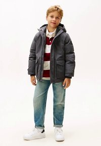 Šedá prošívaná bunda s kapucí, pod ní červenobíle pruhované polo tričko, světle modré džíny a bílé tenisky. Chlapec stojí s rukama v kapsách.