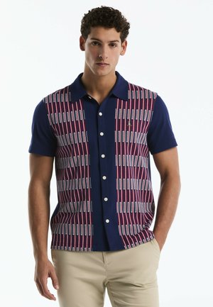 Junger Mann trägt ein kurzärmeliges navyblaues Hemd mit roten und weißen vertikalen Streifen, kombiniert mit beigen Hosen.