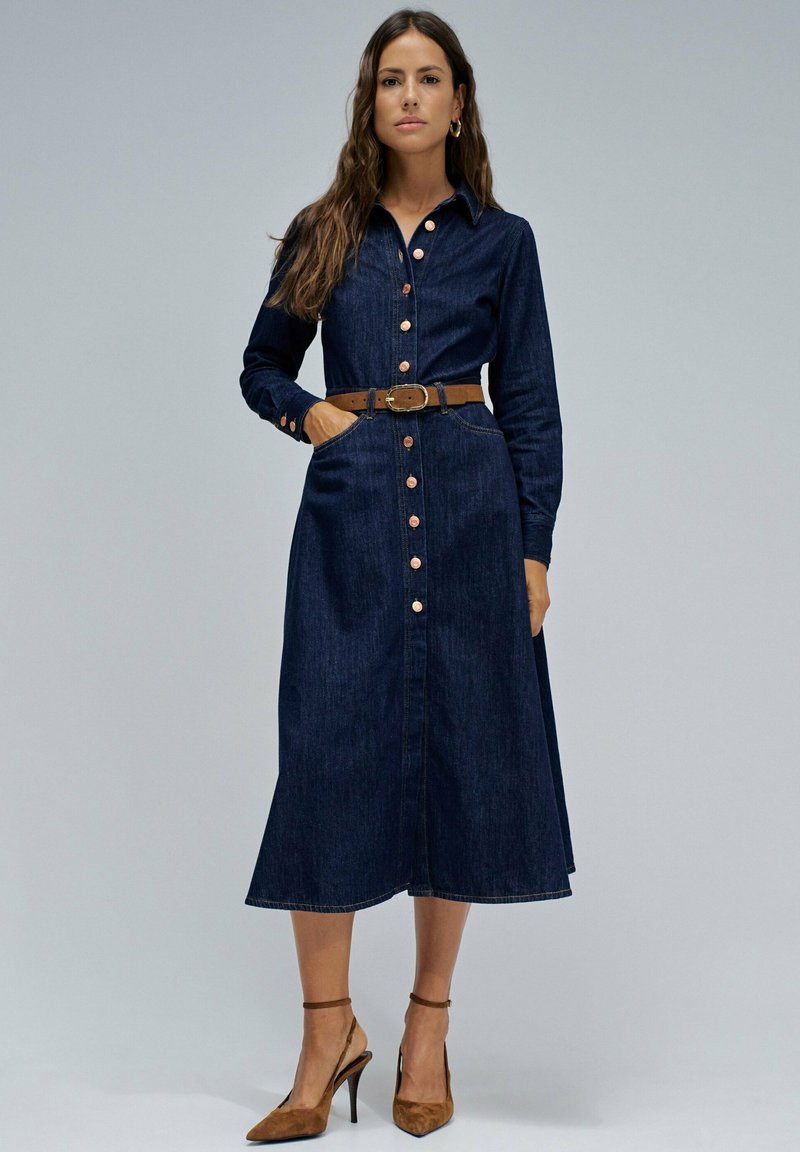Vestido camisa de mezclilla con mangas largas, cierre de botones y bolsillos laterales. Color azul oscuro y viene con un cinturón marrón y zapatos de tacón color beige.