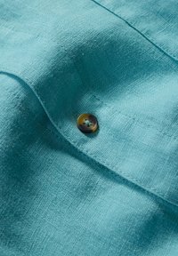 Material de in de culoare albastru deschis cu o textură, având un buton rotund închis la culoare la guler și detalii de cusătură contrastante.