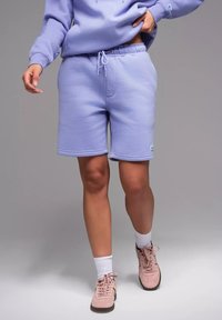 Shorts en polaire lilas avec une taille élastique et des détails de cordon de serrage. Assortis à des baskets roses et des chaussettes blanches de type crew. Texture lisse.
