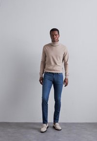 Pulover bej cu guler înalt, blugi skinny albaștri și loafers crema. Textură fină, design ajustat și paletă de culori neutre pe un fundal simplu.