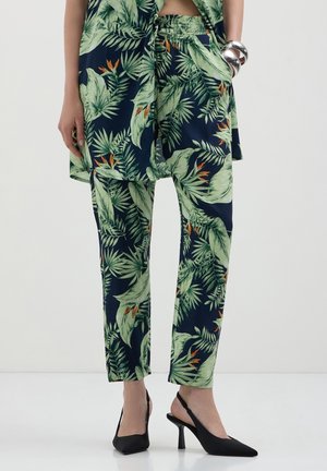 Frau trägt grüne Hose mit tropischem Blattmuster und passendem Oberteil, kombiniert mit schwarzen Slingback-Pumps und silbernen Armbändern.