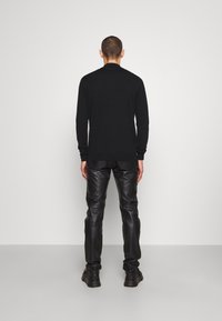 Modèle masculin debout face à un mur, portant un pull noir, un pantalon en cuir noir et des baskets noires sur un fond gris clair uni.