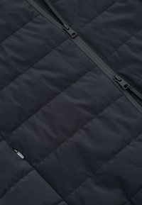 Czarny pikowany materiał z centralnym czarnym zamkiem błyskawicznym i widocznymi suwakami, ukazujący szczegółowe szwy i teksturę.