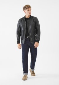 Chaqueta bomber de cuero negro con cuello acanalado, bolsillos con cremallera y un patrón acolchado. Combinada con jeans azules y zapatos beige.