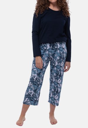 Vrouw draagt een marineblauwe top met lange mouwen en blauwe cropped broek met grote witte en donkere bloemenprint, staat blootsvoets op een witte achtergrond.