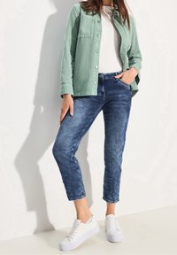 Chemise verte boutonnée portée sur un t-shirt blanc, associée à un jean en denim bleu et des baskets blanches. Caractéristiques : une poche plaquée et des poignets.