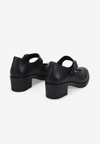 Un par de zapatos Mary Jane de cuero negro con tacones gruesos y correas de hebilla redonda, vistos desde atrás sobre un fondo blanco.