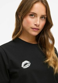 Zwarte t-shirt met een zilveren lippenafdruk op de borst. De stof ziet er glad uit en het shirt heeft een klassieke pasvorm met een ronde hals.