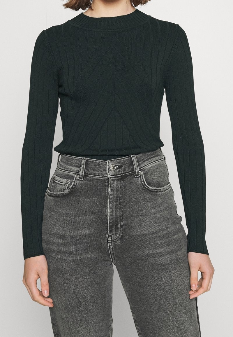 Woman wearing a dark green ribbed long-sleeve sweater tucked into high-waisted faded black jeans with front pockets.