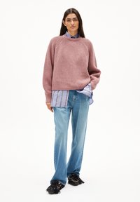 Rosa gerippter Pullover mit Rundhalsausschnitt, über einem blau-weißen, gestreiften Hemd getragen. Hellblaue Schlagjeans und schwarze Fransen Schuhe runden das Outfit ab.