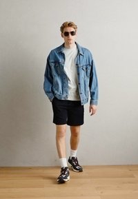 Denimjakke, hvit t-skjorte, svarte shorts. Tilbehør inkluderer mørke solbriller og mønstrede joggesko. Bakgrunn med tregulv.